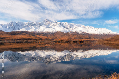 Beautiful autumn mountain lake and mountains. Reflection of snowy peaks and red coniferous taiga on the surface of a mountain lake. Russia. Altai Republic. Panoramic view.