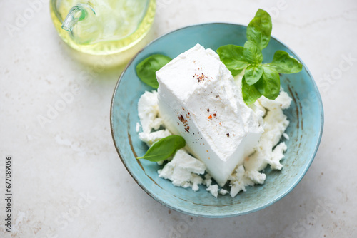 Turquoise bowl with a block of feta cheese and green basil on a light-beige stone background, horizontal shot, elevated view