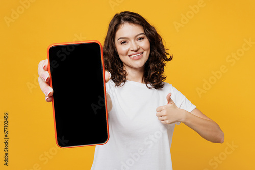 Young woman wear white blank t-shirt casual clothes hold in hand use blank screen workspace area mobile cell phone show thumb up isolated on plain yellow orange background studio. Lifestyle concept.