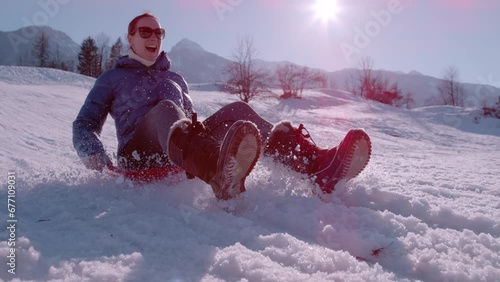 SLOW MOTION, CLOSE UP: Happy young woman enjoys sledging down the snowy hill. While sledding on plastic snow sledge, she is smiling and excited. Fun filled outdoor winter activities in alpine valleys. photo