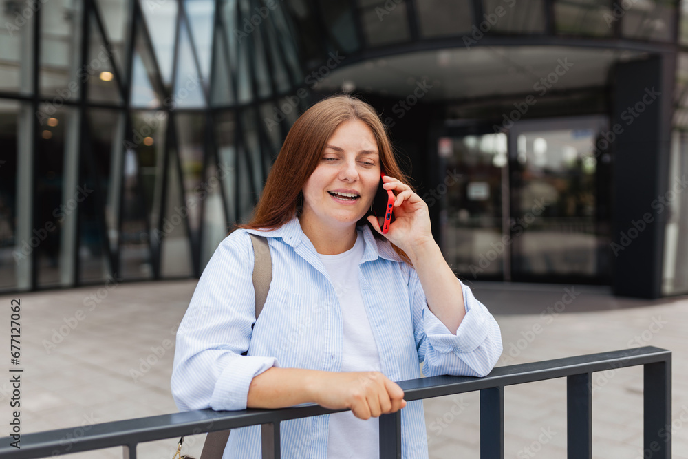 Fototapeta premium Young happy redhead businesswoman making phone call over mobile phone in the city. Middle age woman talking on smartphone, walking city urban street at business office center. Manager work outside