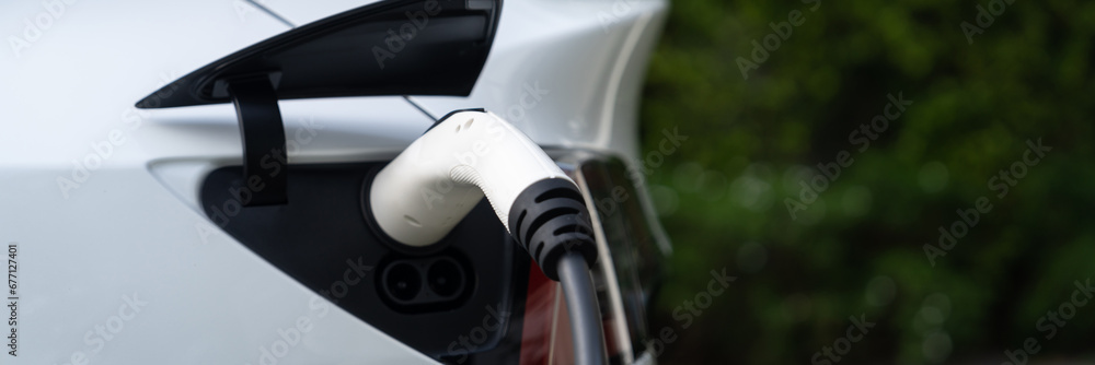 Closeup EV charger plug handle attached to electric vehicle port, recharging battery from charging station. Modern designed EV car and clean energy sustainability for better future.Panorama Synchronos