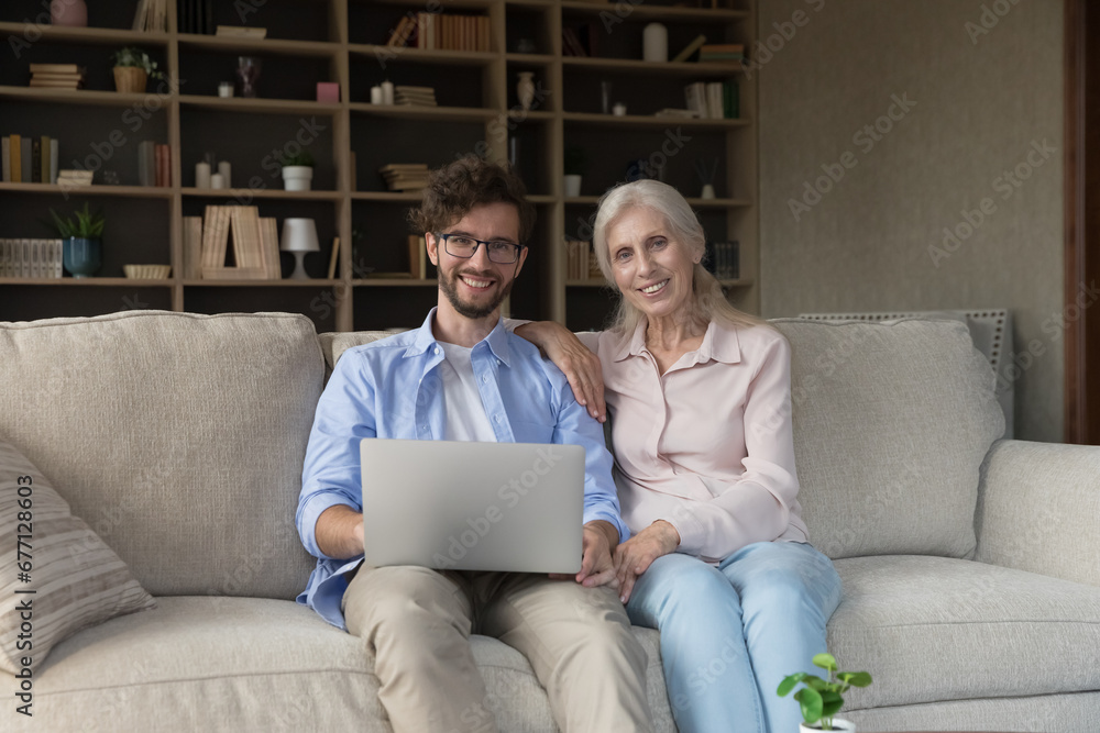 Smiling millennial man spend time with retired mother at home, sit on sofa with laptop, look at camera, young adult son helps with computer usage to senior mom. Two generations family use modern tech