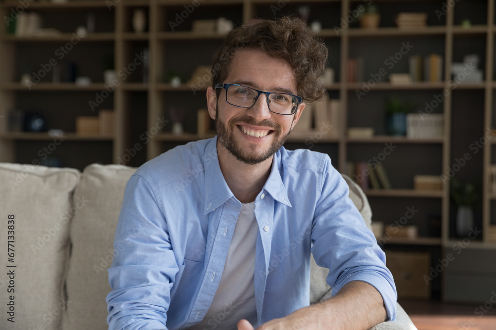 Fototapeta premium Happy millennial 35s handsome man sit on sofa posing, staring at camera. Bearded guy in eyeglasses spend time at home, enjoy carefree relaxation at own or rented apartment. Leisure, homeowner portrait