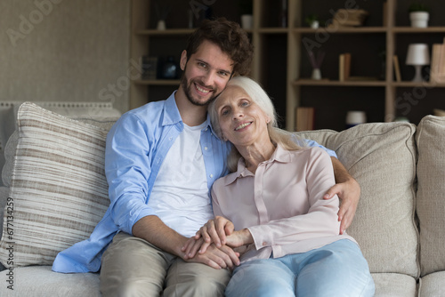 Smiling grownup son sit on cozy sofa with older mom, enjoy leisure time together in living room pose, looking at camera, handsome man embrace senior mother express love and care. Family relationship