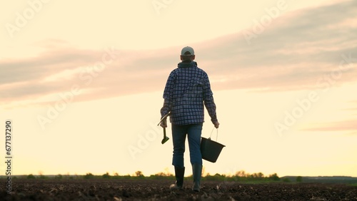 Agricultural industry. Farmer in boots walks across field. Farmer working in field, sunset. Agricultural business concept. Owner of farm with shovel, bucket in hand, go along plowed field. Plantation