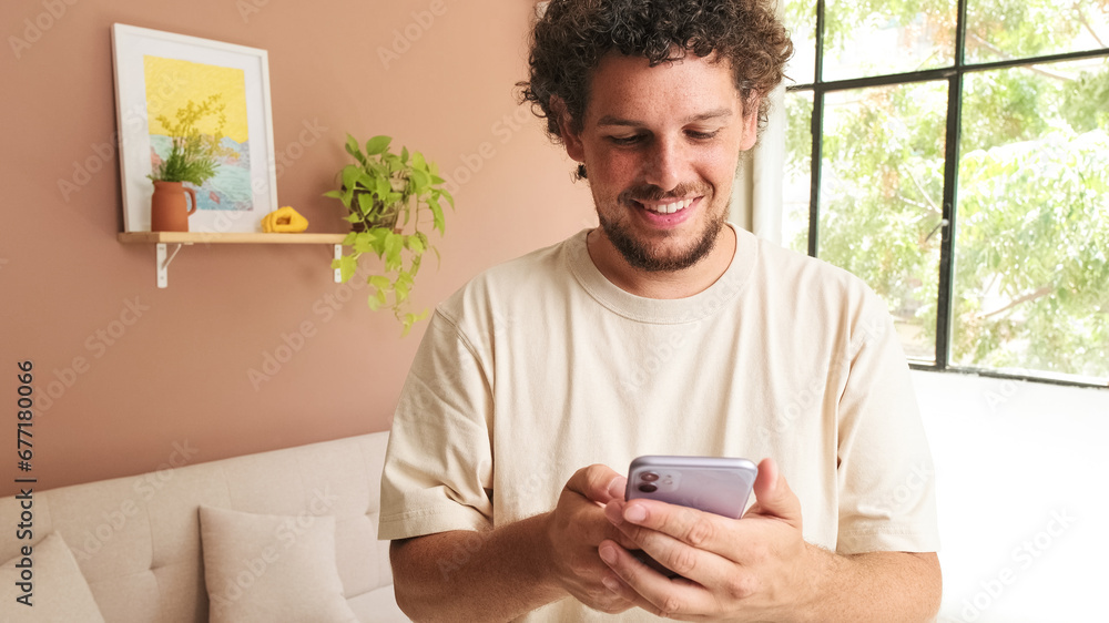 Smiling young man with curly hair and beard using mobile phone while standing in cozy living room Smiling young man with curly hair and beard using mobile phone while standing in cozy living room