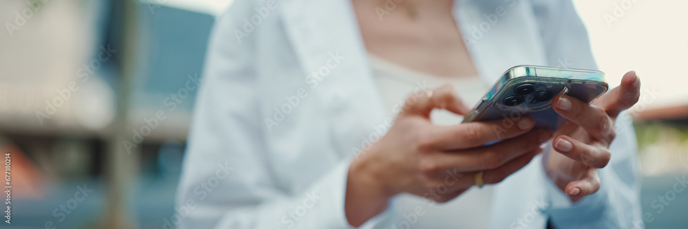 Closeup of young woman holds smartphone in her hands and scrolls through the news feed. Close-up of girl hand uses mobile phone outdoors - obrazy, fototapety, plakaty 