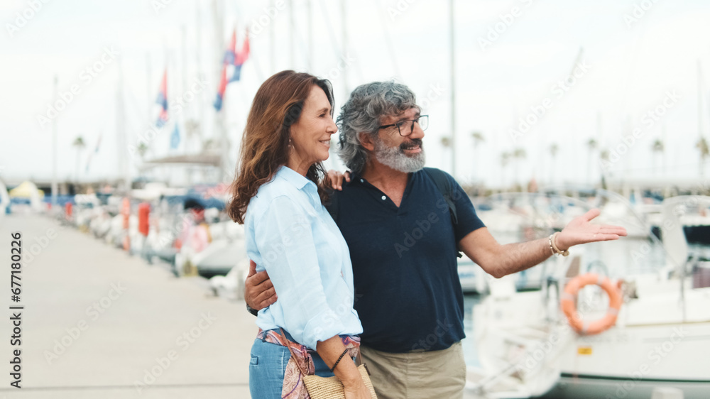 Obraz premium Elderly couple in love are happy on walk together, they are planning sea voyage while standing in the port on yacht background