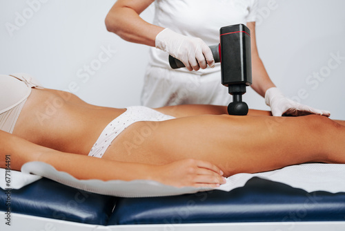 Young sporty caucasian woman having body massage using percussion pistol at beauty relax center. Electric massager pistol for special muscle treatment. Sports and pain recovery concept.