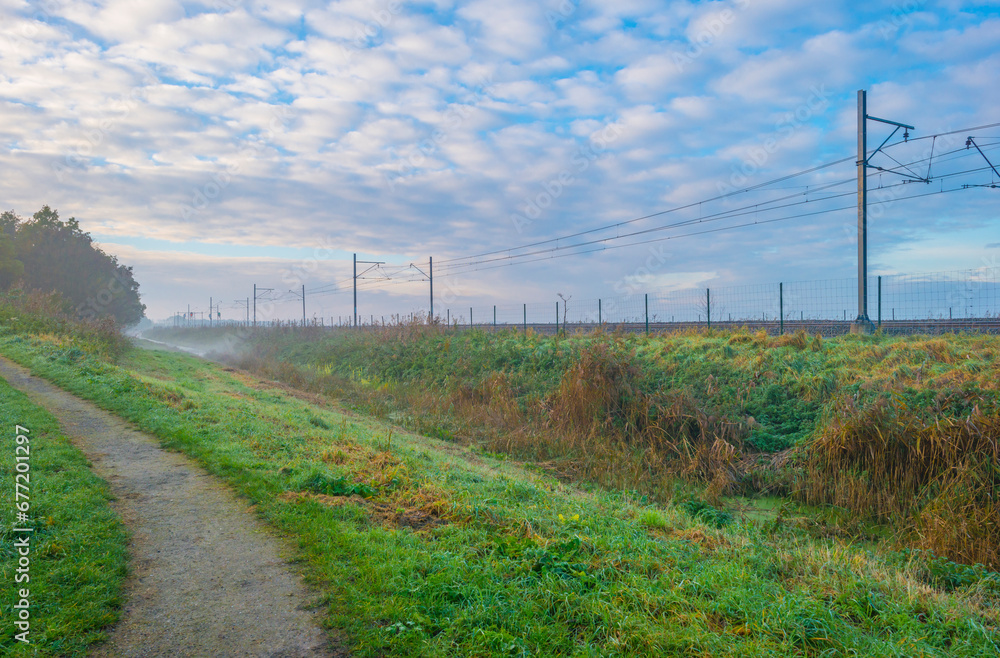 Railway track in a green field with reed in wetland beneath a blue cloudy sky in winter, Almere, Flevoland, The Netherlands, November 12, 2023