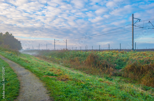 Railway track in a green field with reed in wetland beneath a blue cloudy sky in winter, Almere, Flevoland, The Netherlands, November 12, 2023