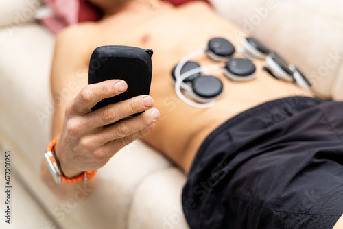 An unrecognizable man uses an electro stimulator on his abs on the couch at home to reduce muscle tension and help relaxation. Improve your muscle strength  optimize your rehabilitation.