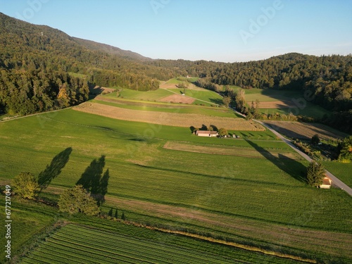 Aerial horizontal image of a green field with a forest on a hill in the distance and a clear sky