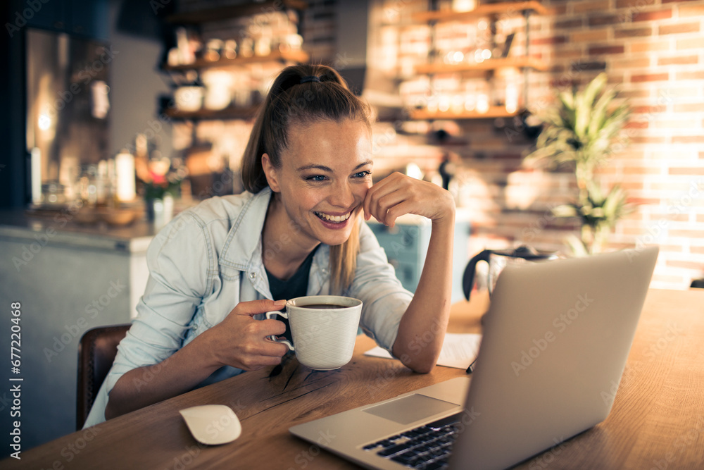 Smiling young woman with coffee cup sitting at desk with laptop - obrazy, fototapety, plakaty 