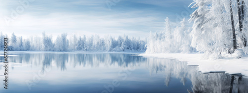 A serene winter landscape with a snow-covered forest reflecting on the surface of a tranquil, partly frozen lake under a clear blue sky.