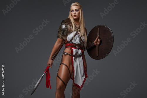 Viking woman, clad in fantasy armor with bare thighs. She confidently wields a shield and spear on a gray backdrop