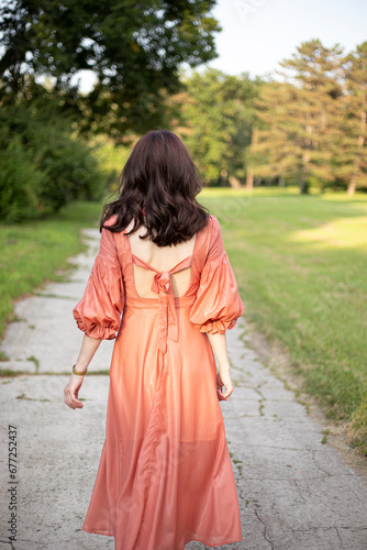 Back view of a  brunette girl in dress posing in the park, tree background.

