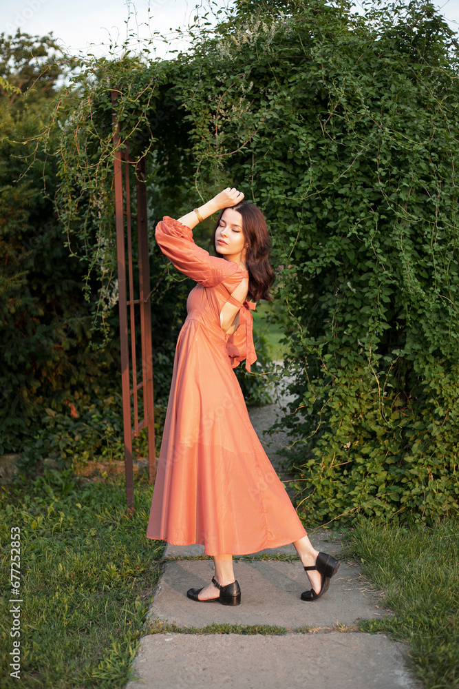 Full length of a beautiful brunette girl in orange dress posing in the park, green background.
Full length of a beautiful brunette girl in orange dress posing in the park, green background.