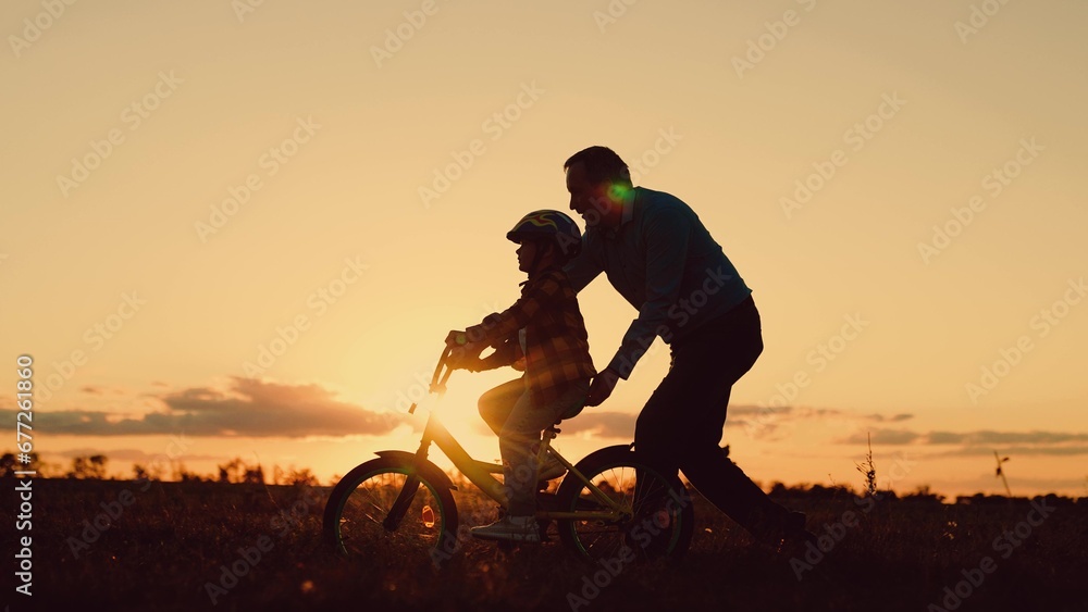 Father helps his daughter ride bike. Father teaches child wearing safety helmet to ride bicycle in park. Child rides bicycle. Kid, dad play together, sunset. Child dream learns to ride bicycle. Family - obrazy, fototapety, plakaty 