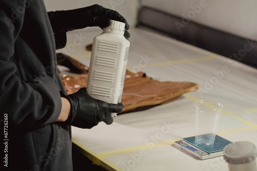 A woman in black protective gloves takes a white bottle with a primer in the workshop. Unscrews the bottle cap. There is a leather bag in the background