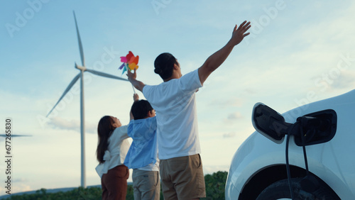 Concept of progressive happy family enjoying their time at wind farm with electric vehicle. Electric vehicle driven by clean renewable energy from wind turbine generator for charging station.