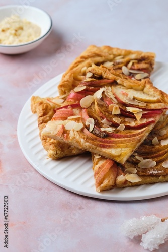 Sliced open apple pie on puff pastry with apple slices, sprinkled with almond petals, on a white ceramic board on a pink marble background.