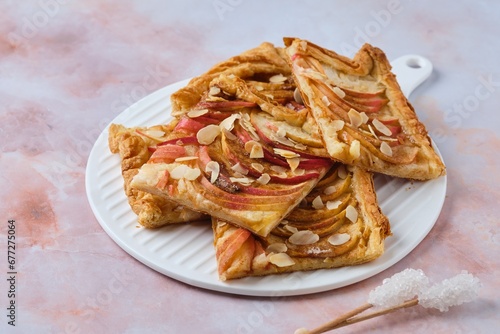 Sliced open apple pie on puff pastry with apple slices, sprinkled with almond petals, on a white ceramic board on a pink marble background.