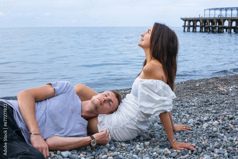 A guy lies on a girl's lap on a pebble beach by the sea