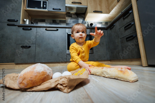 A cute 1-year-old boy is sitting in the kitchen with fresh bread. Child with bread on the floor. The child eats bread photo