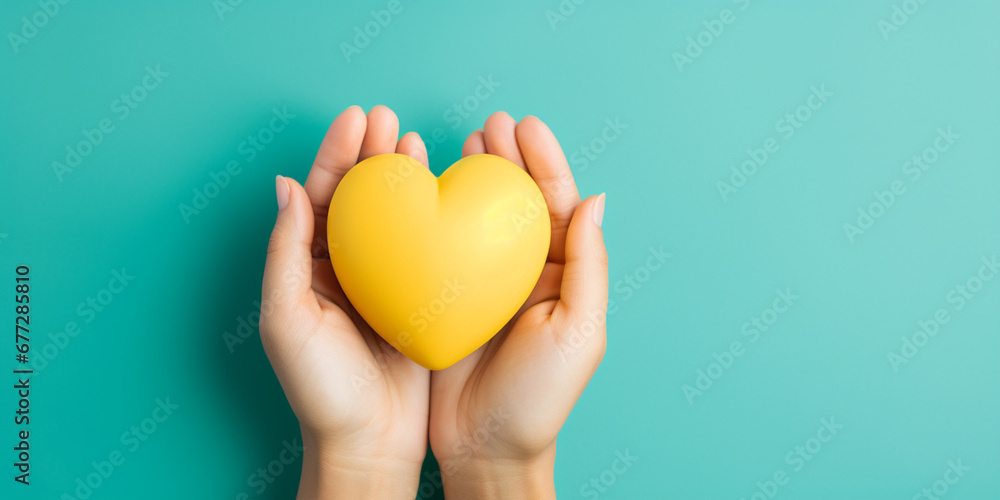 The woman hand is holding a yellow heart on teal blue background - obrazy, fototapety, plakaty 