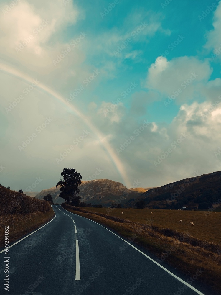 Long straight road surrounded by green grass and mountains under the beautiful rainbow and clouds