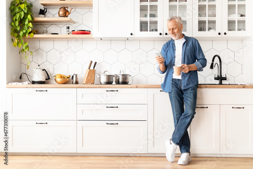 Mobile Communication. Smiling Senior Man Relaxing In Kitchen With Smartphone And Coffee