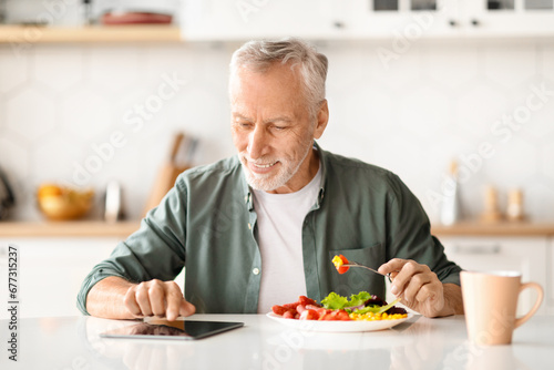 Cheerful Senior Man With Digital Tablet Reading News During Breakfast In Kitchen
