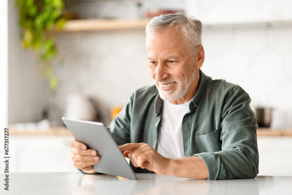 Happy Senior Man Using Digital Tablet While Relaxing At Table In Kitchen Happy Senior Man Using Digital Tablet While Relaxing At Table In Kitchen