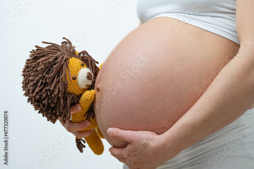 Close-up of a pregnant mother in whote cloths holding a stuffed lion and her hand on her bare round belly, expecting the baby. Last month of pregnancy - week 39. White background. Bright shot. photo