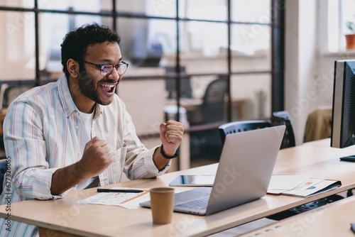 Cheerful Hispanic man celebrating success with laptop in office