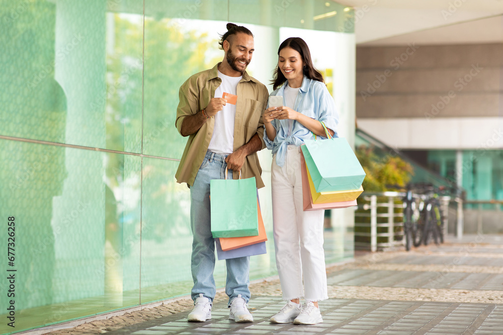 Cheerful millennial european couple with bags enjoy shopping app on smartphone, credit card in mall