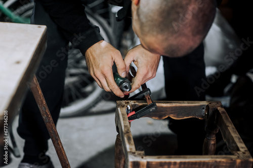 An adult male professional worker, woodworker, carpenter glues a chair, repairs, restores furniture indoors, workshop, clamping wooden products with a clamp. Photography, work concept, portrait.