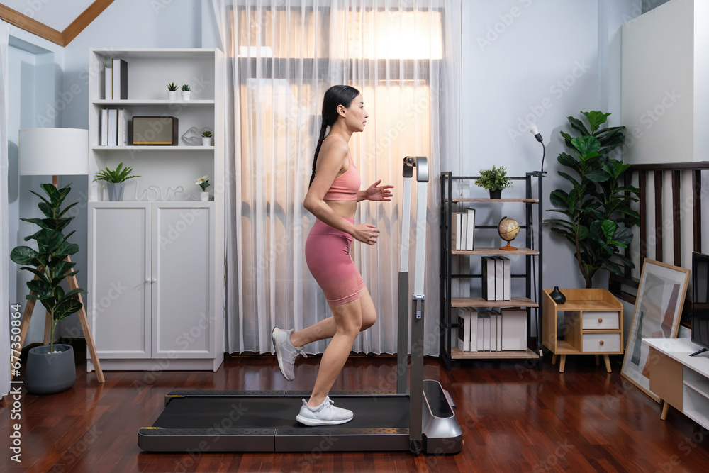 Full length side view of energetic and strong athletic asian woman running running machine at home. Pursuit of fit physique and commitment to healthy lifestyle with home workout and training. Vigorous - obrazy, fototapety, plakaty 