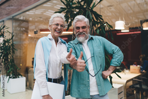 Vibrant senior Caucasian couple in a co-working space giving thumbs up, possibly celebrating a successful business venture. The woman is in her 70s, with white hair, holding a laptop