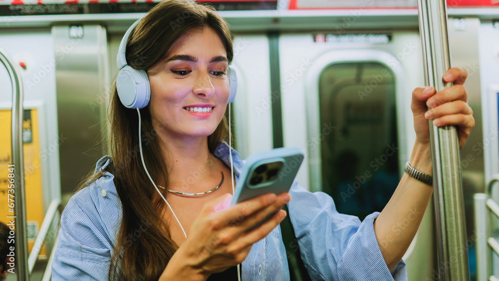 Female chatting online in mobile phone messenger watch video in metro. Young girl listening music in headphones and hold smartphone in subway car travel underground use wireless internet connection. - obrazy, fototapety, plakaty 