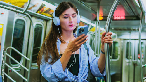 Young adorable charming Caucasian woman looking at smartphone texting with serious expression. Beautiful stylish girl commuting in underground listening to music with headphones.