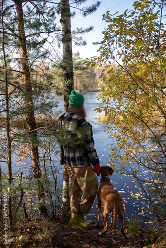 Pet owner with hound dog magyar vizsla standing on steep, looking at pond, listening to silence, breathing fresh air. Female spending pastime in Scandinavian autumn pine forest with beloved pet 