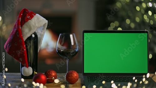 Glass and bottle of red wine with laptop computer on table in Christmas decorated home. Christmas tree and lights in winter home with fireplace. Green screen for web shop ads and vinery holiday offer.