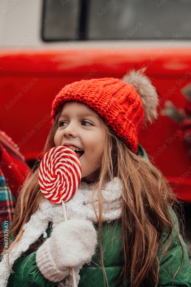 Little girl in red knitted hat with Xmas candy cane in hands outdoor