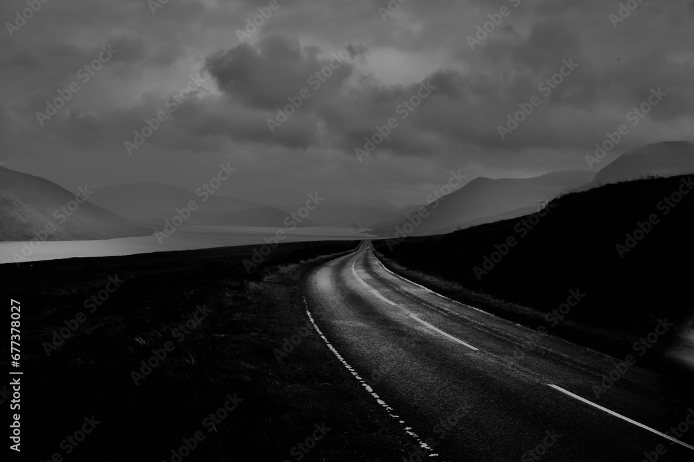 Greyscale of a road surrounded by a field to rocky smooth mountains
