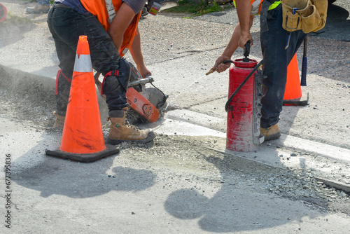 Two construction workers wearing belts cut a hole in a concrete sidewalk using a mechanical saw. An orange safety cone is on the road and sidewalk. Concrete dust is blowing around the street. 
 photo