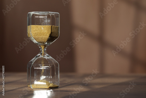 Hourglass with flowing gold sand on wooden table against brown background, space for text