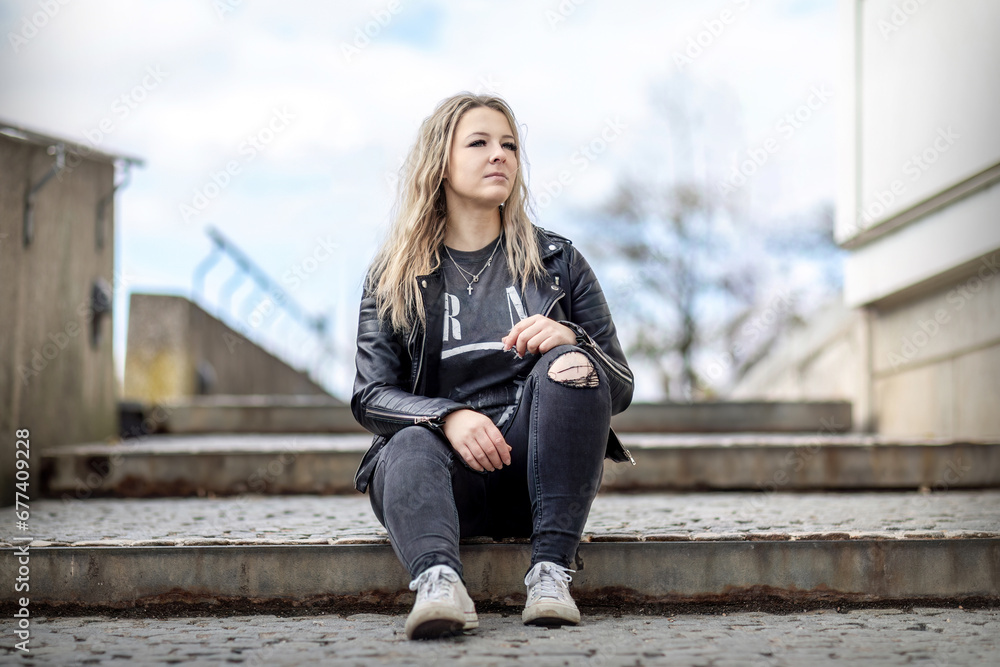 A young blonde woman sitting on stairs in a city and looks thoughtful, streetstyle portrait